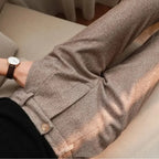 Close-up of a person wearing textured taupe-brown trousers with a buttoned waist detail and a black top, seated indoors on a light beige sofa.