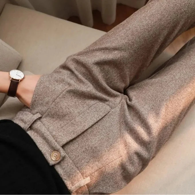Close-up of a person wearing textured taupe-brown trousers with a buttoned waist detail and a black top, seated indoors on a light beige sofa.