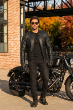 A man wearing a black leather jacket, black t-shirt, black jeans, and black boots stands in front of a black cruiser motorcycle on a paved street. He is wearing sunglasses and black leather gloves, with a brick building and autumn trees in the background.