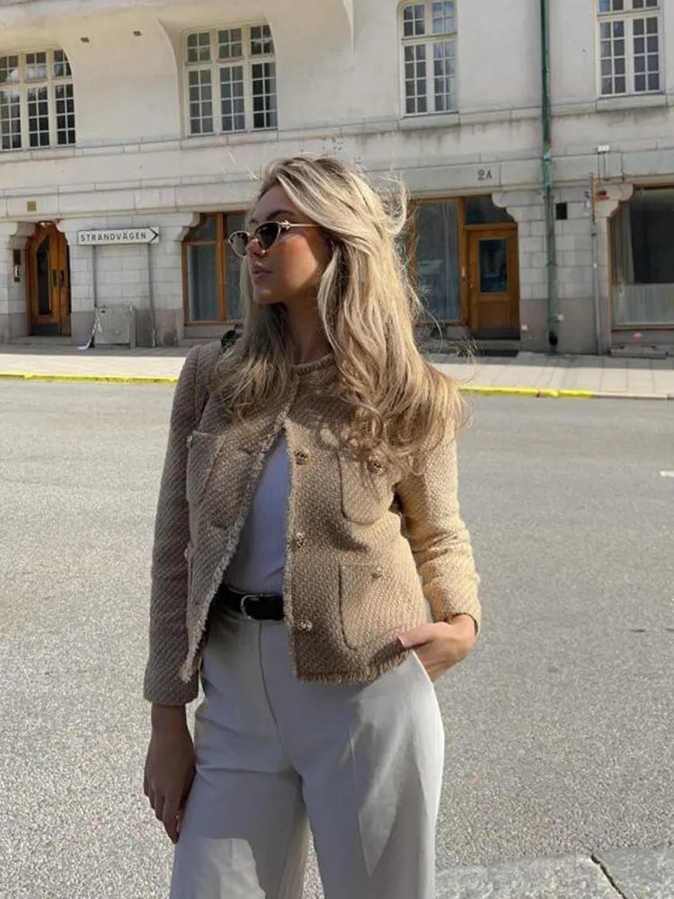 Woman in a beige jacket and light pants standing on a street with buildings in the background