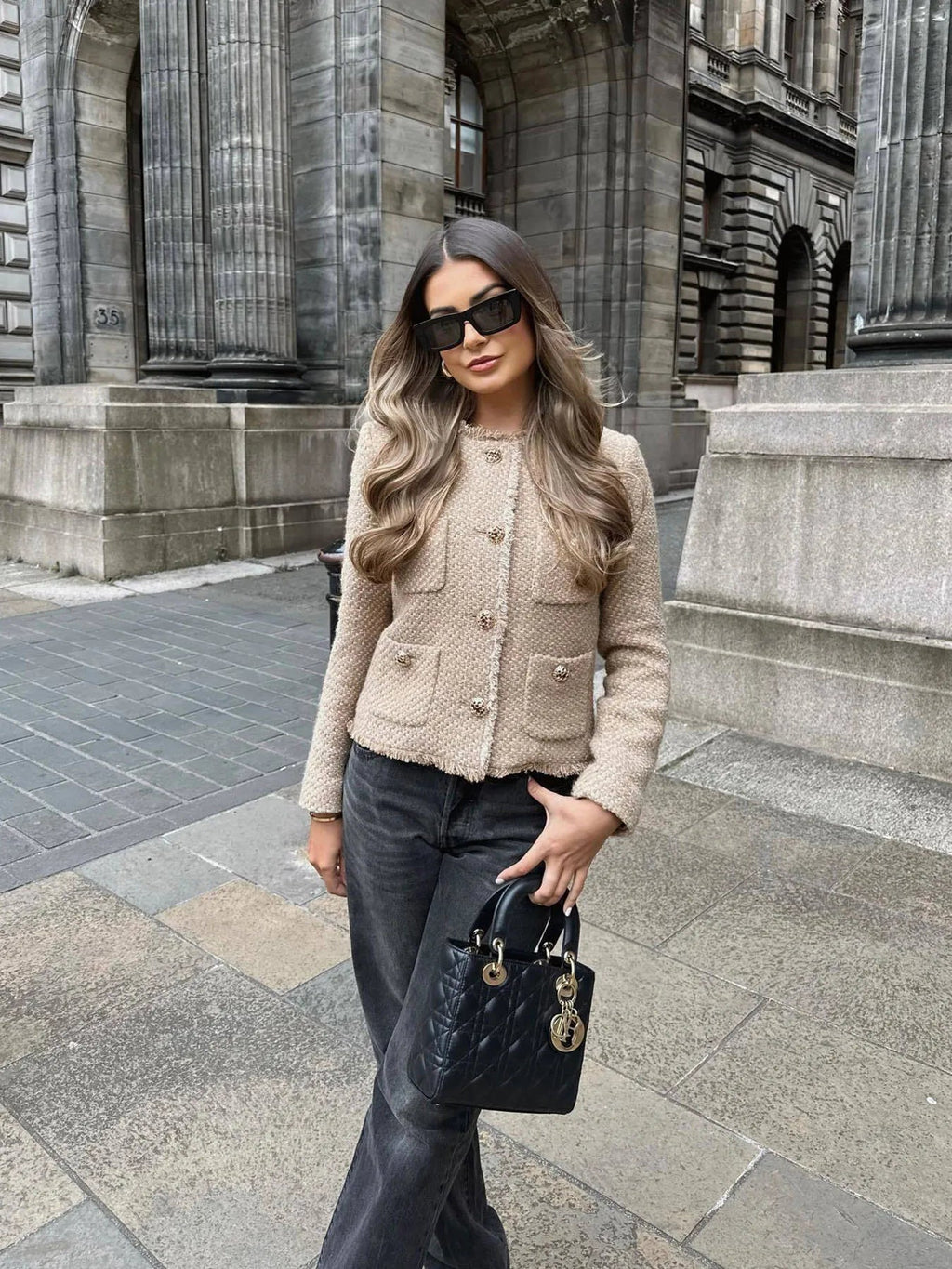 Woman in a beige jacket and dark pants standing in front of a building.