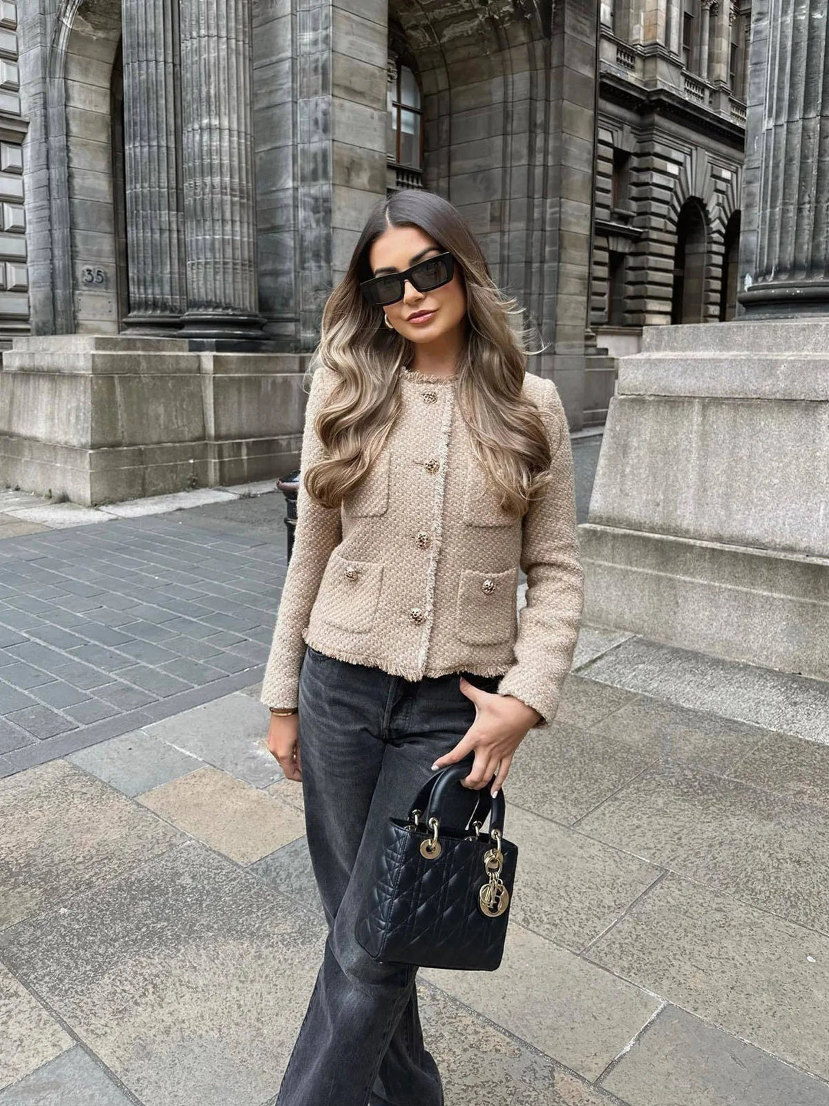 Woman in a beige jacket and dark pants standing in front of a building.