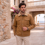 A man wearing a tan wool-blend jacket with a beige sherpa collar, standing in a stone courtyard.