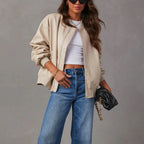 Woman wearing a beige jacket, white top, and blue jeans against a gray background