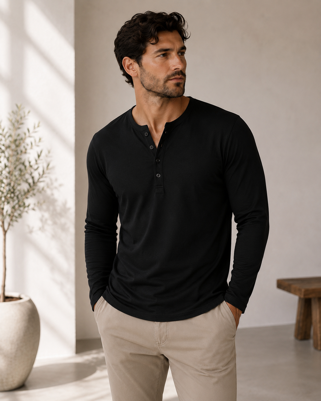 A man wearing a black long-sleeve henley shirt and beige pants, standing in a brightly lit room with a potted plant and wooden bench in the background.