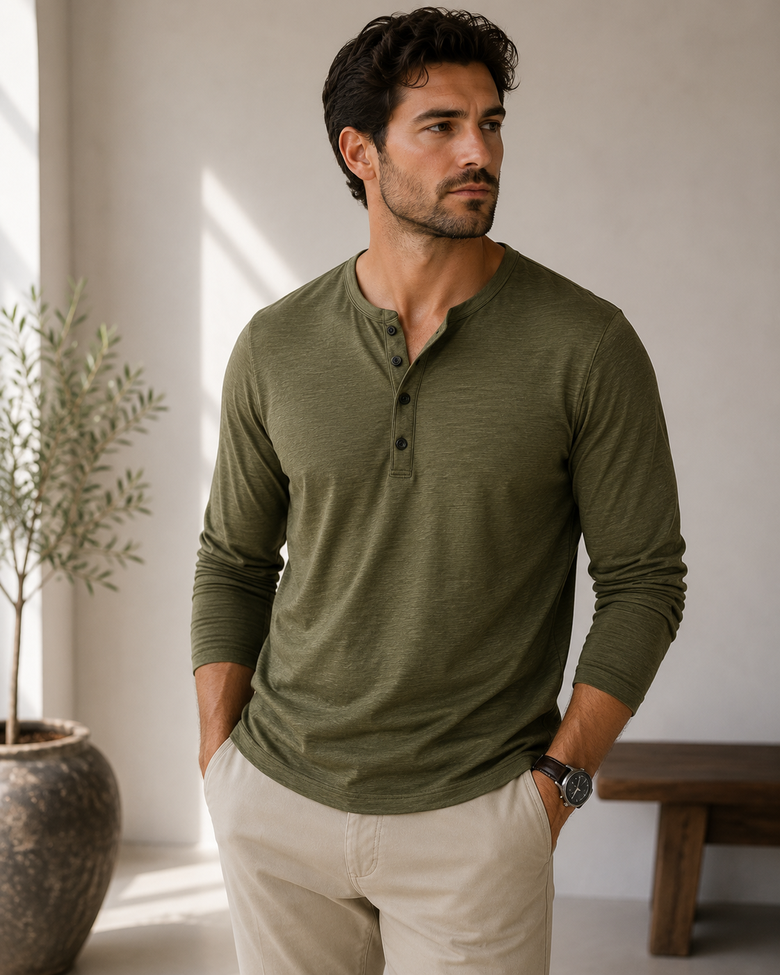 A man wearing a long-sleeve olive green henley shirt and beige pants, standing in a bright indoor setting with a potted plant and wooden bench in the background.