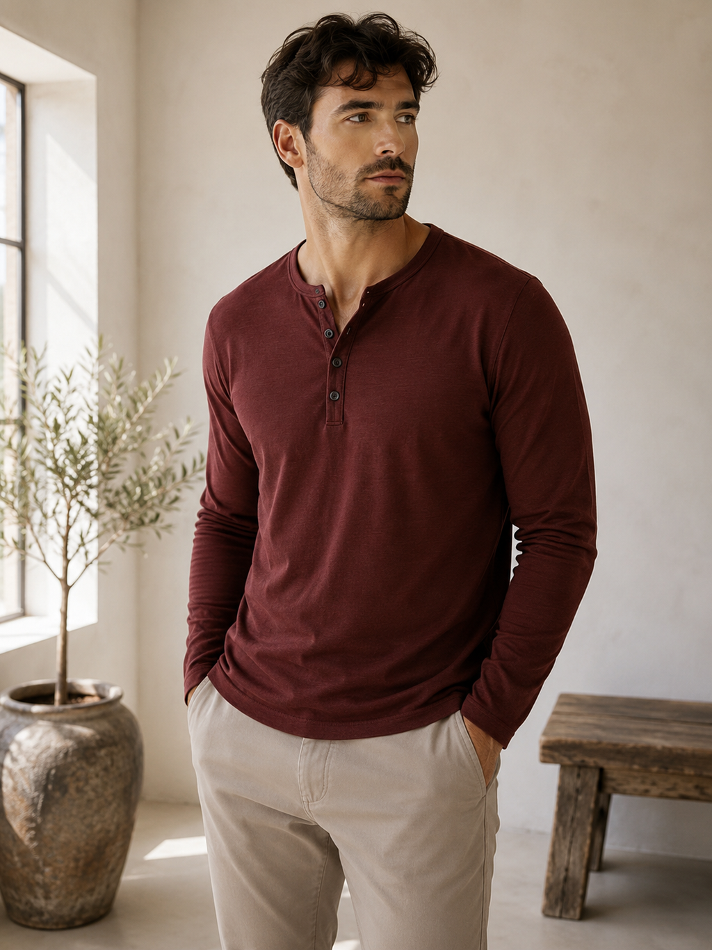 A man wearing a burgundy long-sleeve henley shirt and beige trousers, standing in a bright, neutral-toned room with a potted plant and wooden bench in the background.