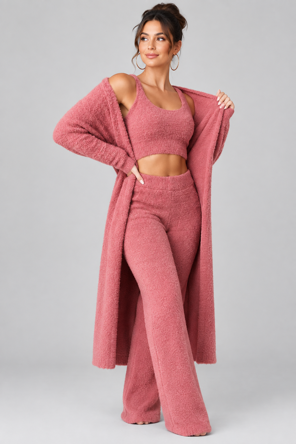 A woman modeling a matching fuzzy three-piece loungewear set in dusty rose pink, featuring a long open-front cardigan, a cropped tank top, and high-waisted flared pants, photographed against a light gray studio background.