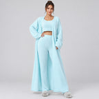 A model wearing a light blue three-piece loungewear set consisting of a cropped tank top, matching high-waisted wide-leg pants, and a long open-front cardigan, styled with beige slippers against a plain white background.