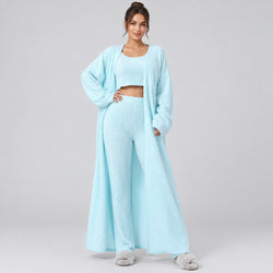 A model wearing a light blue three-piece loungewear set consisting of a cropped tank top, matching high-waisted wide-leg pants, and a long open-front cardigan, styled with beige slippers against a plain white background.