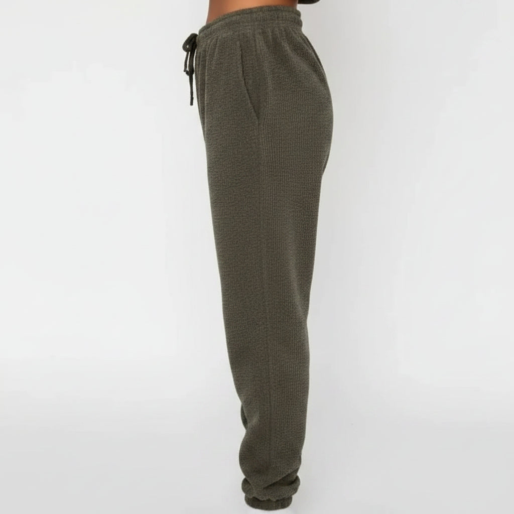 A person shown from the waist down wearing dark olive green textured jogger sweatpants with a drawstring waist and white sneakers, against a plain white studio background.