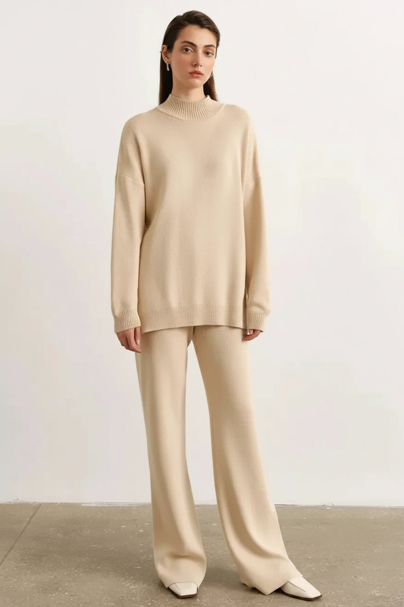 A woman modeling a matching beige knit two-piece set consisting of a long-sleeve mock-neck sweater and wide-leg trousers, standing against a minimalist off-white wall and concrete floor.