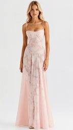 A woman modeling a floor-length light pink gown with thin straps, structured bodice, and a flowing skirt with tonal floral embroidery, photographed against a plain light gray studio background.