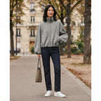 A woman stands on a city sidewalk wearing a light gray fuzzy quarter-zip pullover, dark blue jeans, and white sneakers, holding a taupe tote bag. The background shows blurred trees and a building.