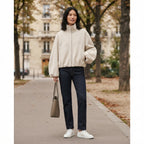 A woman stands on a city sidewalk wearing a cream-colored zip-up fleece jacket, dark blue jeans, and white sneakers, holding a taupe tote bag. The background shows blurred trees and a building.