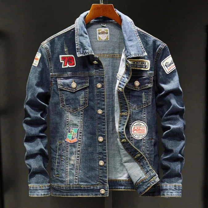 A blue denim jacket with multiple colorful patches and a silver-tone snap-button front, hanging on a wooden hanger against a dark background.