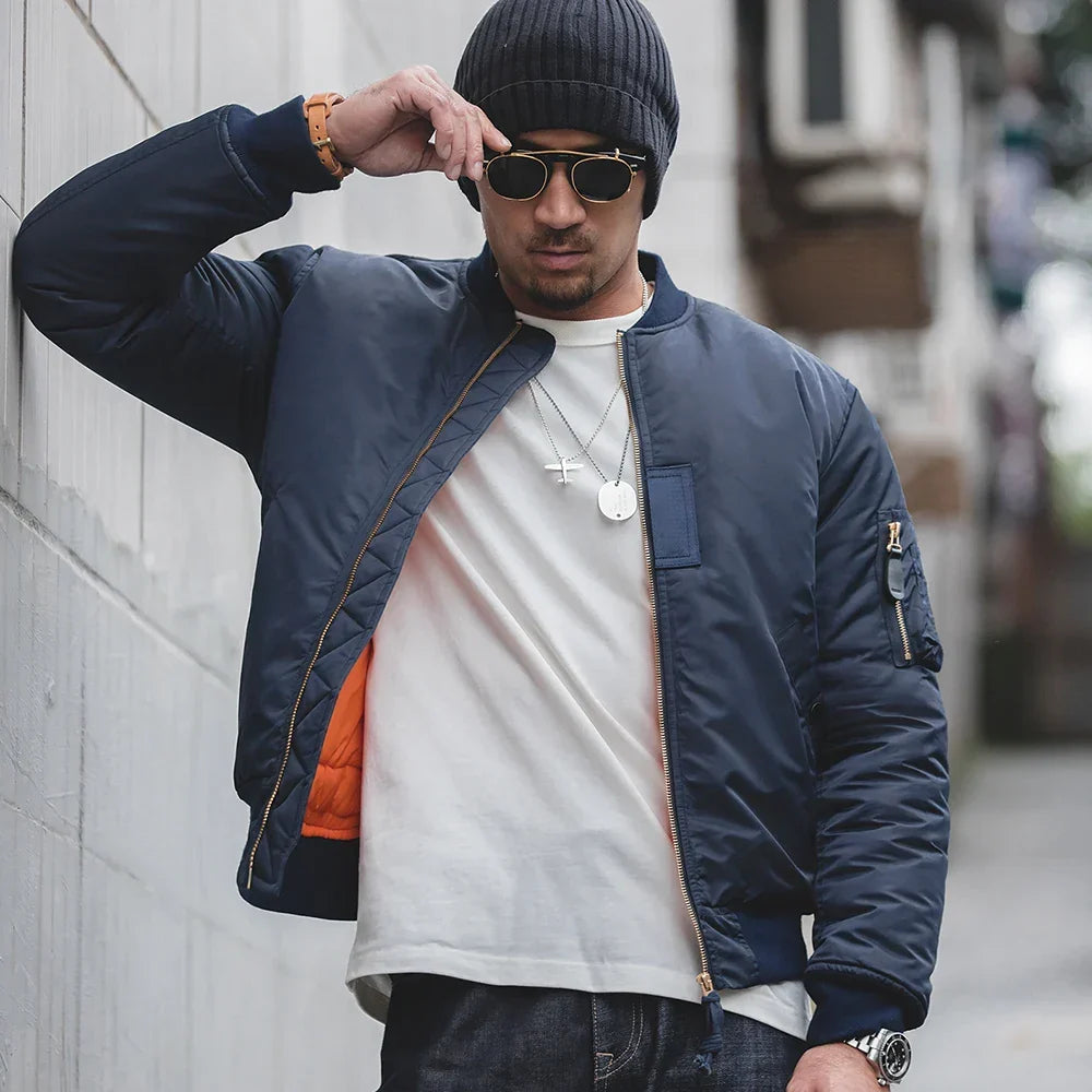 Man wearing a navy bomber jacket, white t-shirt, and dark sunglasses on a city street.