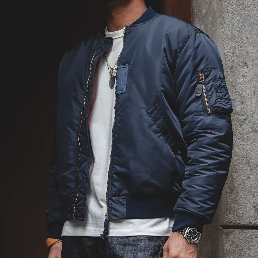 Person wearing a blue bomber jacket with a white shirt underneath, standing against a dark background.