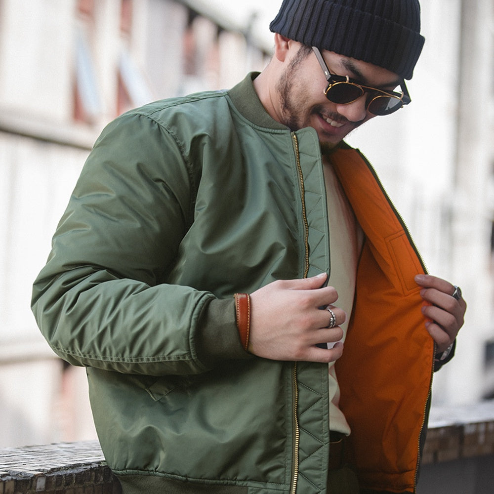 Man wearing a green jacket and black beanie with sunglasses, holding an orange bag.