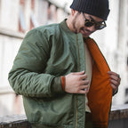 Man wearing a green jacket and black beanie with sunglasses, holding an orange bag.