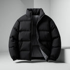 A black puffer jacket with a high stand-up collar and front zipper, displayed on a white geometric platform against a light gray studio background.