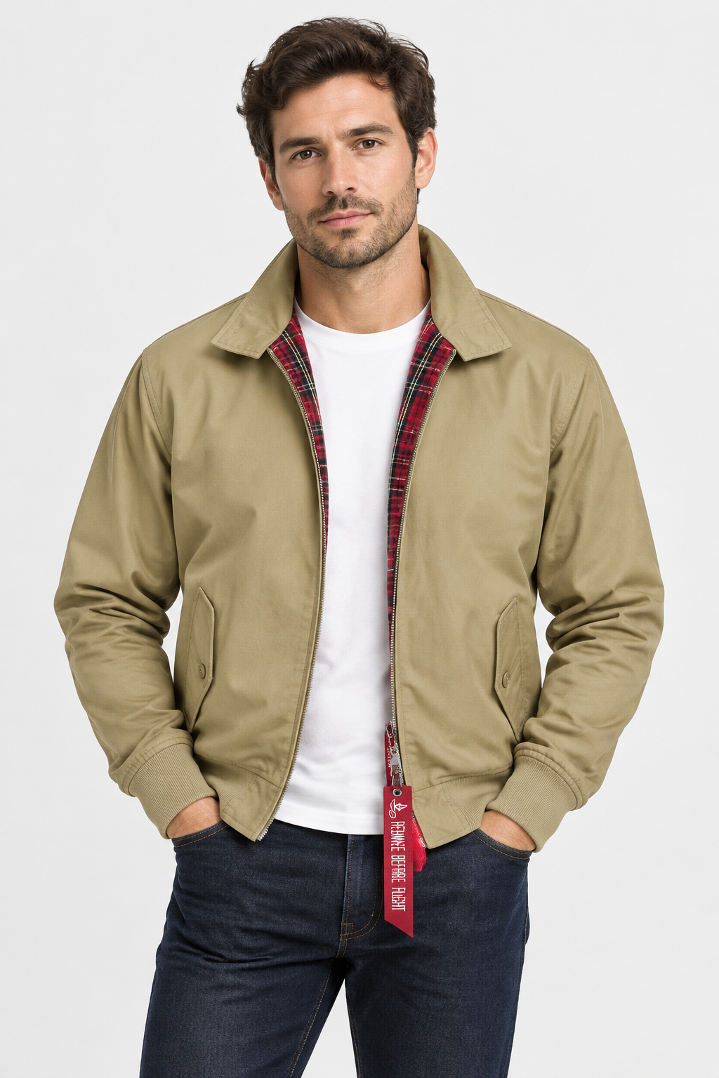 A man wearing a tan zip-up overshirt with a red plaid inner lining over a white t-shirt and dark blue jeans, standing against a light gray studio background.