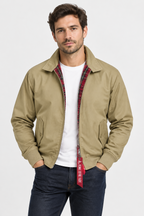 A man wearing a tan zip-up overshirt with a red plaid inner lining over a white t-shirt and dark blue jeans, standing against a light gray studio background.