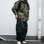 Person wearing a green jacket and black pants standing against a white wall.