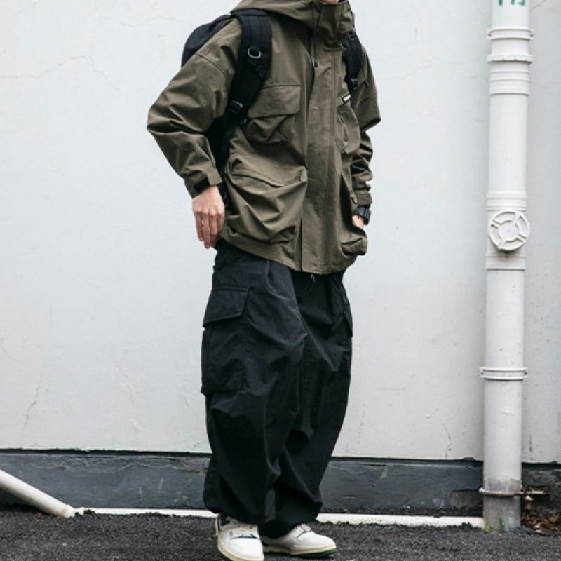 Person wearing a green jacket and black pants standing against a white wall.