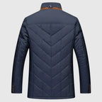 A men's quilted jacket shown from the back, featuring a deep V-pattern on the back panel and sleeves, a brown collar accent, and button details on the cuffs, set against a light gray background.