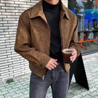 Person wearing a brown suede jacket holding a glass of dark liquid on a sidewalk.