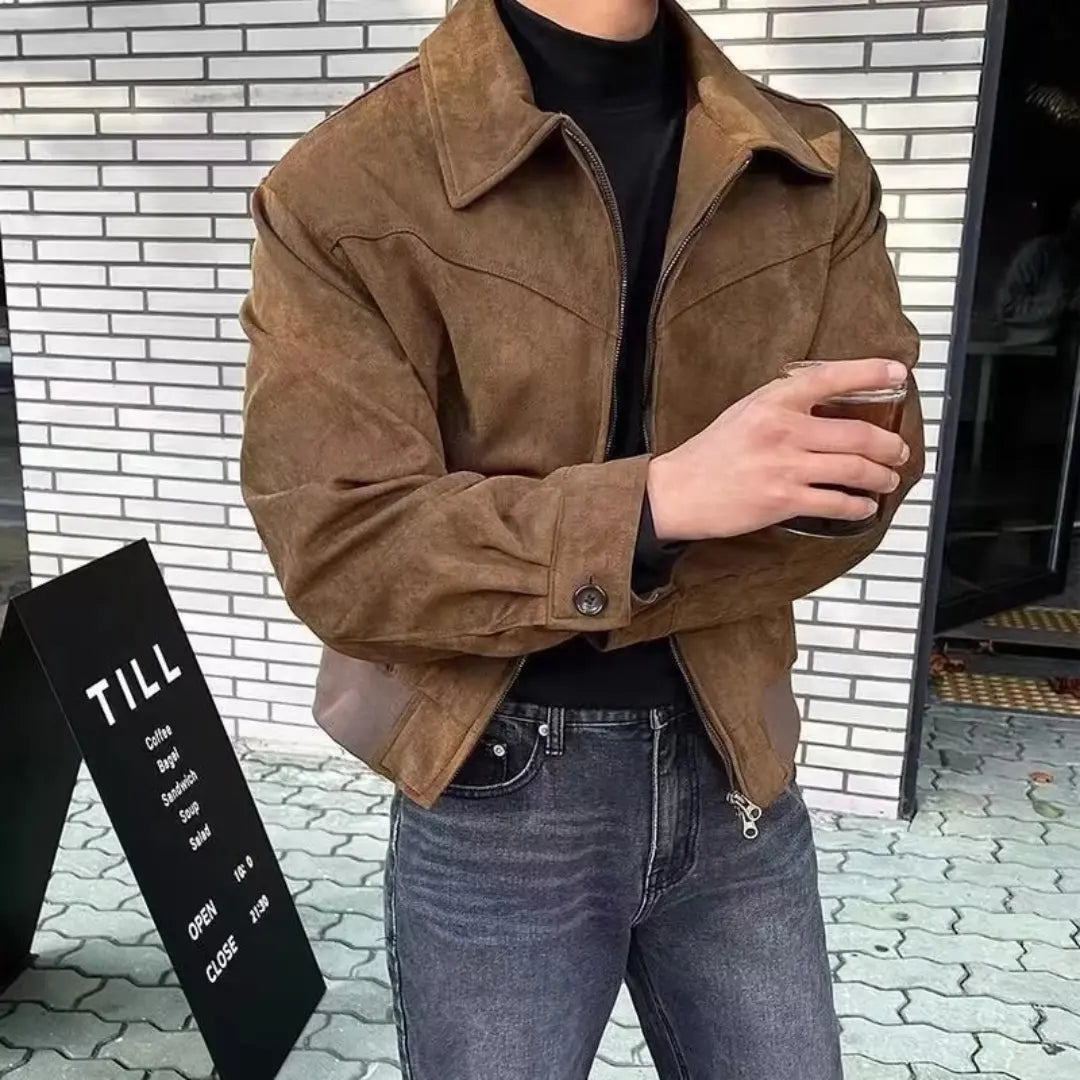 Person wearing a brown jacket and holding a drink against a brick wall.