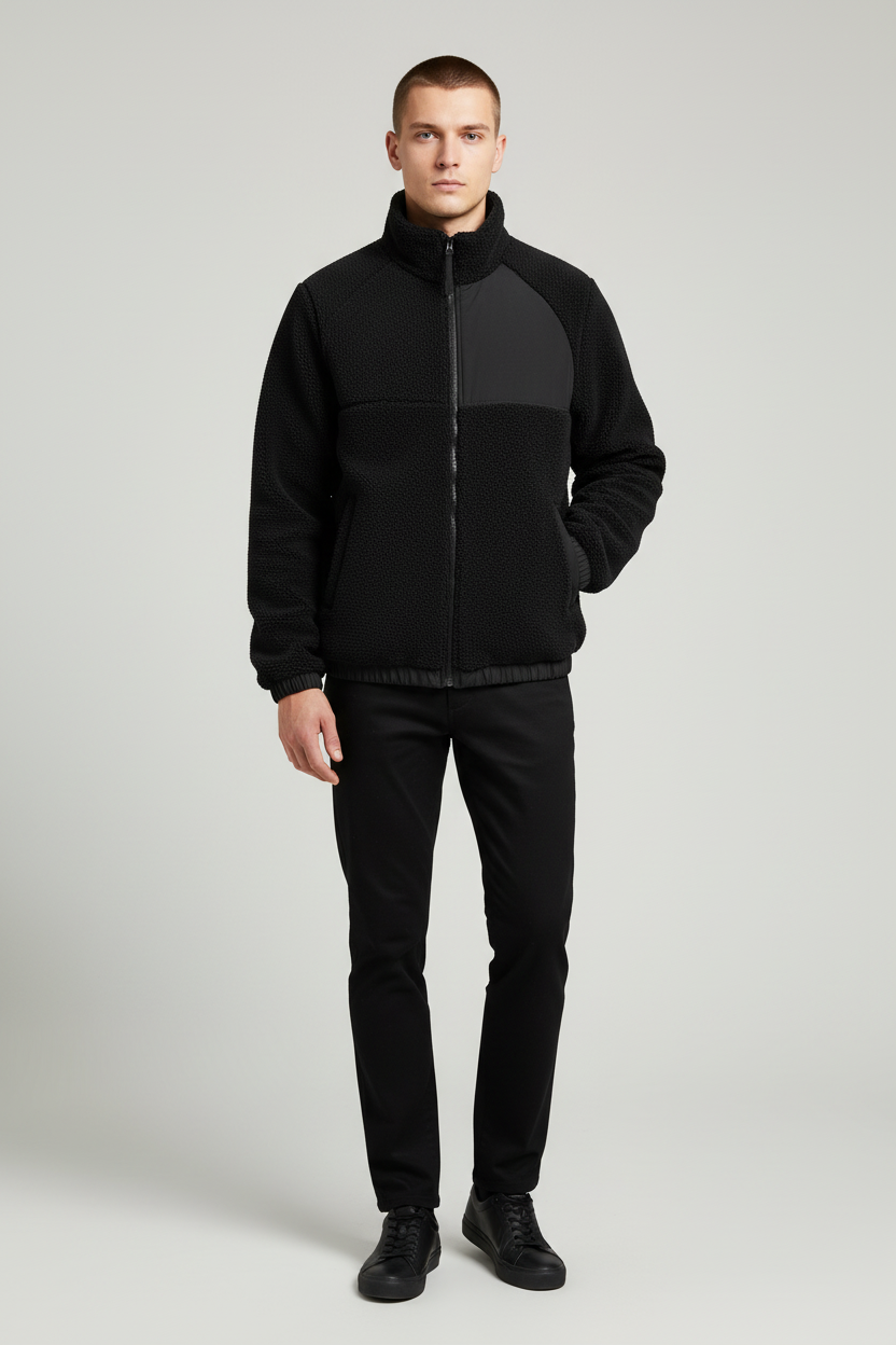 A man modeling a black color-block fleece jacket with a high collar and front zipper, paired with black pants and black shoes, against a light gray studio background.