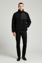 A man modeling a black color-block fleece jacket with a high collar and front zipper, paired with black pants and black shoes, against a light gray studio background.