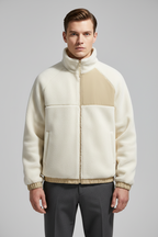 A man wearing a cream and beige color-blocked fleece jacket with a high collar and zip-up front, paired with dark gray trousers, against a solid gray studio background.
