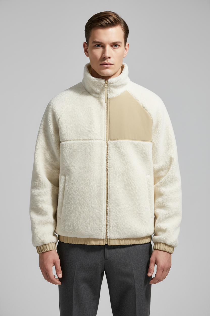 A man wearing a cream and beige color-blocked fleece jacket with a high collar and zip-up front, paired with dark gray trousers, against a solid gray studio background.