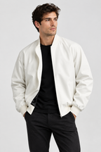 A man wearing a white bomber jacket over a black t-shirt and black pants, standing against a light gray studio background.