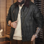 A bearded man wearing a dark waxed cotton jacket with a brown corduroy collar over a cream cable-knit sweater, standing in a rustic indoor setting.
