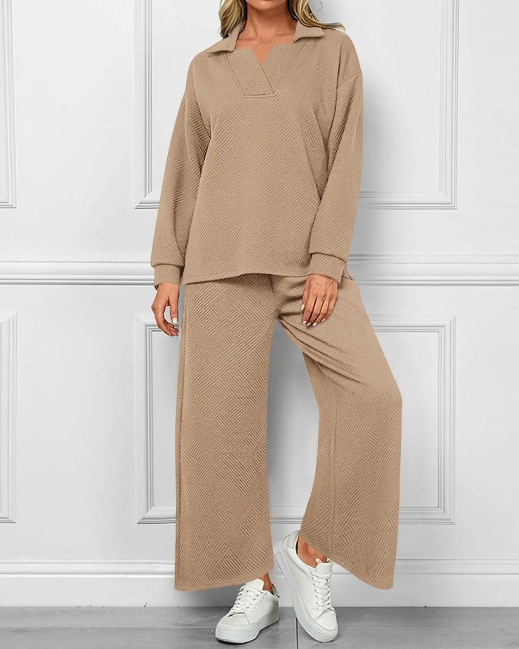 A woman modeling a matching two-piece lounge set in a warm beige color, featuring a long-sleeve collared top and wide-leg pants with a subtle textured pattern, paired with white sneakers against a white paneled wall.