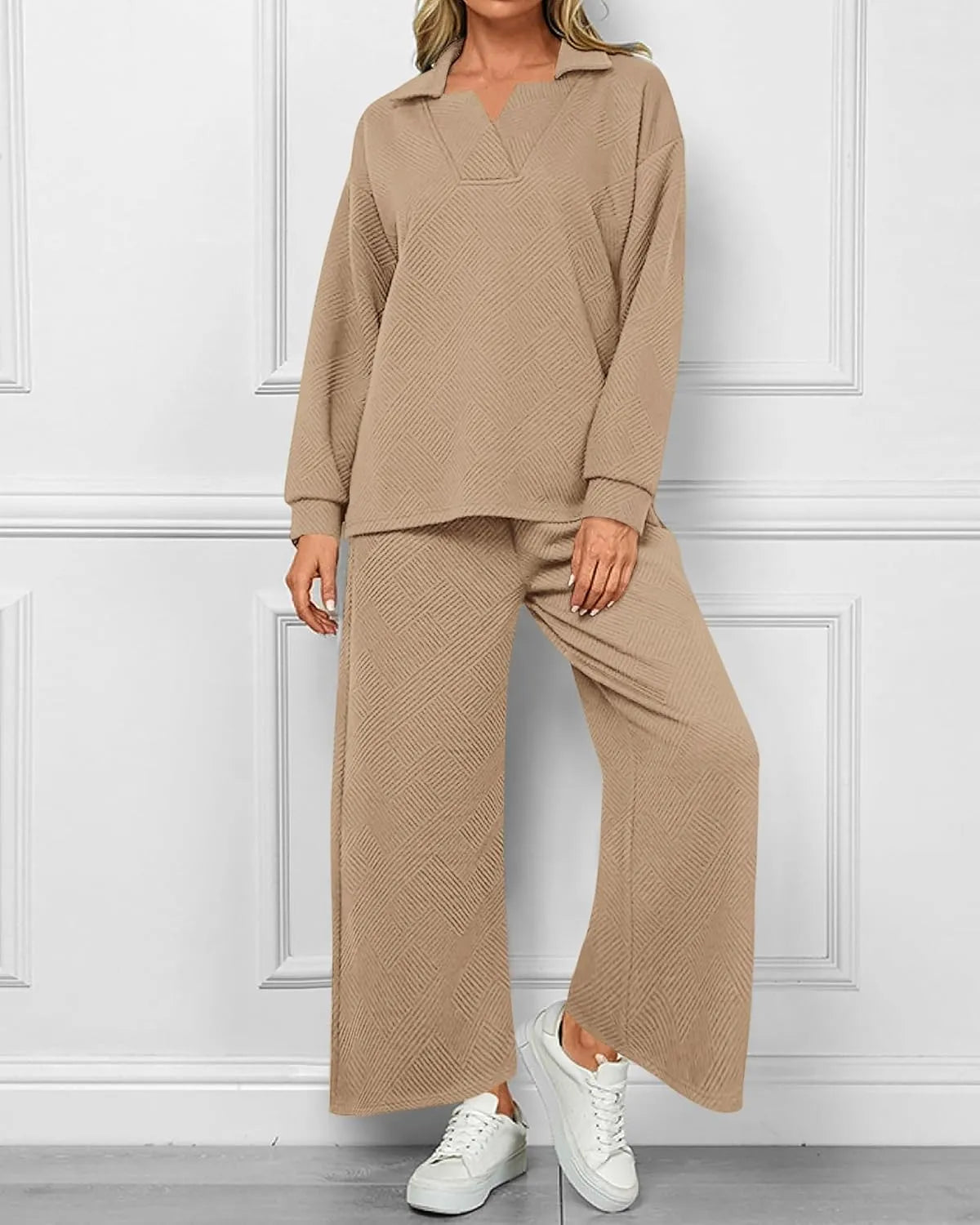 A woman modeling a matching two-piece lounge set in a warm beige color, featuring a long-sleeve collared top and wide-leg pants with a subtle textured pattern, paired with white sneakers against a white paneled wall.