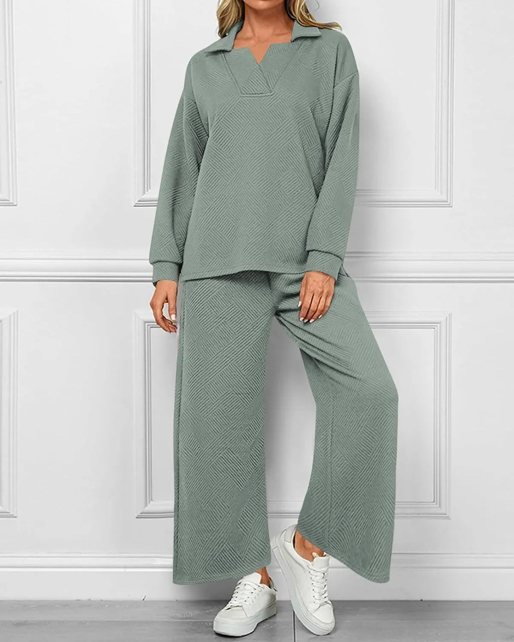 A woman models a matching two-piece lounge set consisting of a long-sleeve collared top and wide-leg pants in a textured sage green fabric, paired with white sneakers against a white paneled wall.