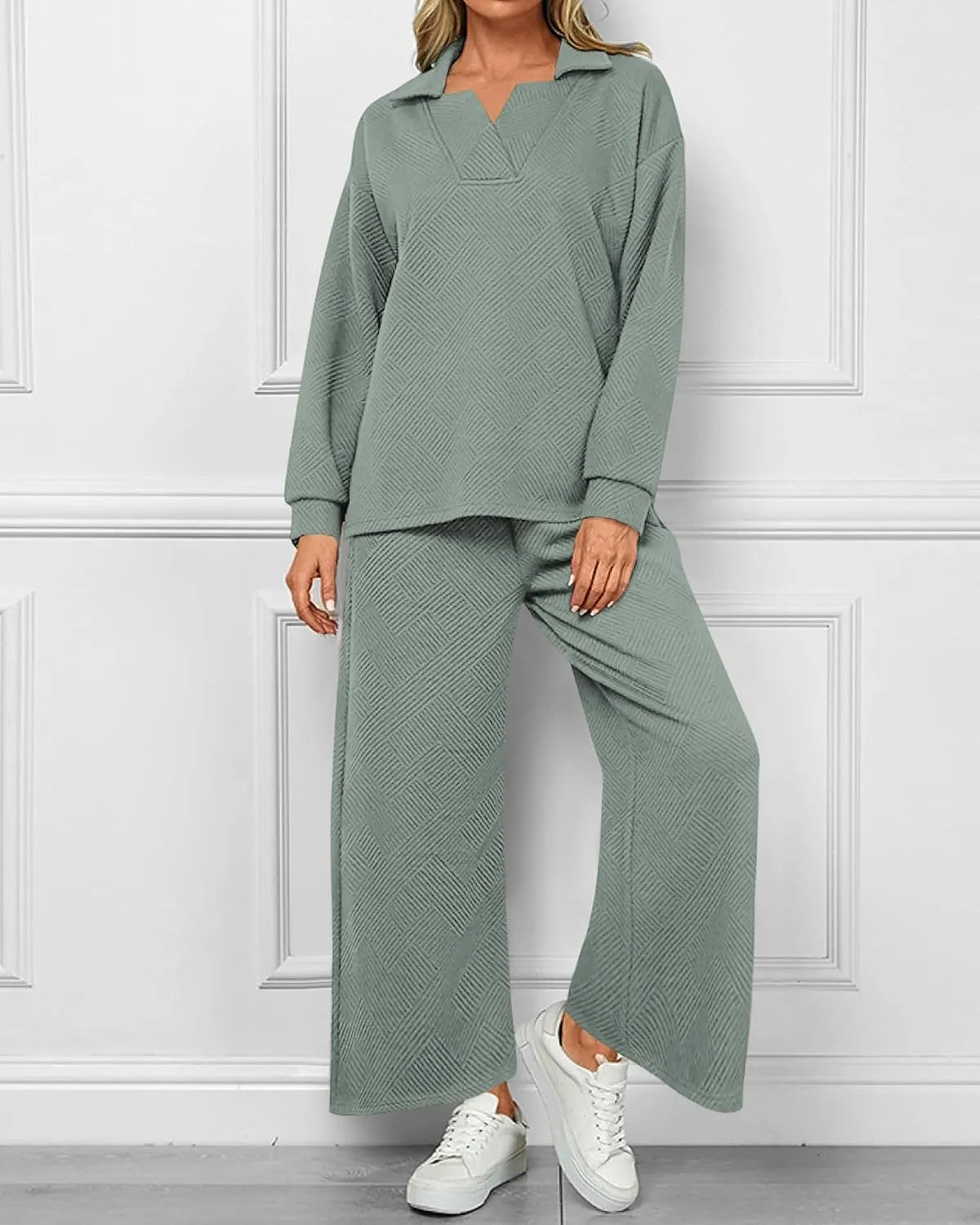 A woman models a matching two-piece lounge set consisting of a long-sleeve collared top and wide-leg pants in a textured sage green fabric, paired with white sneakers against a white paneled wall.