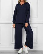 A woman modeling a matching two-piece lounge set in dark navy blue, featuring a long-sleeve collared top and wide-leg pants, paired with white sneakers against a white paneled wall.