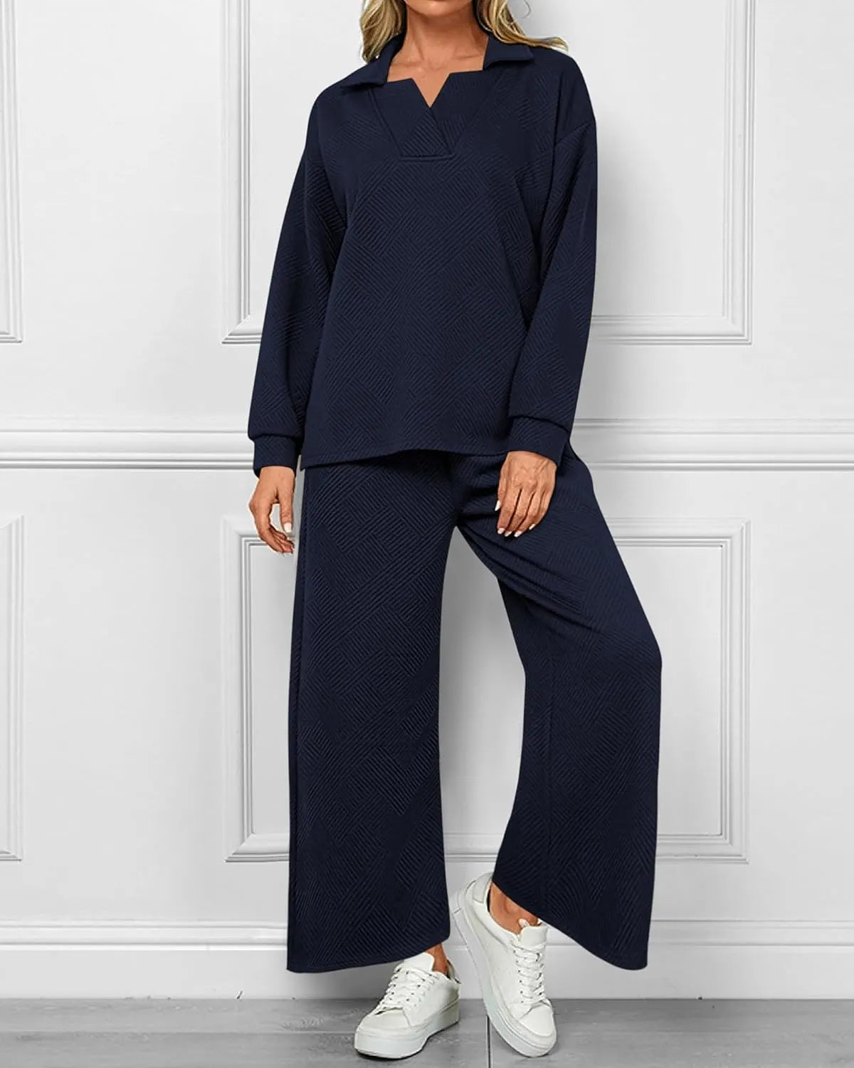A woman modeling a matching two-piece lounge set in dark navy blue, featuring a long-sleeve collared top and wide-leg pants, paired with white sneakers against a white paneled wall.