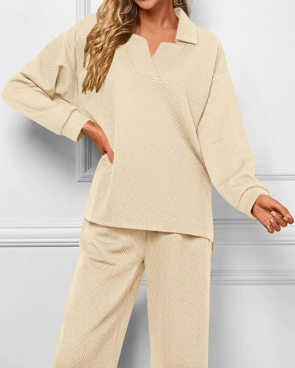A woman wearing a matching beige knit lounge set with a long-sleeve collared top and wide-leg pants, shown against a light gray paneled wall.