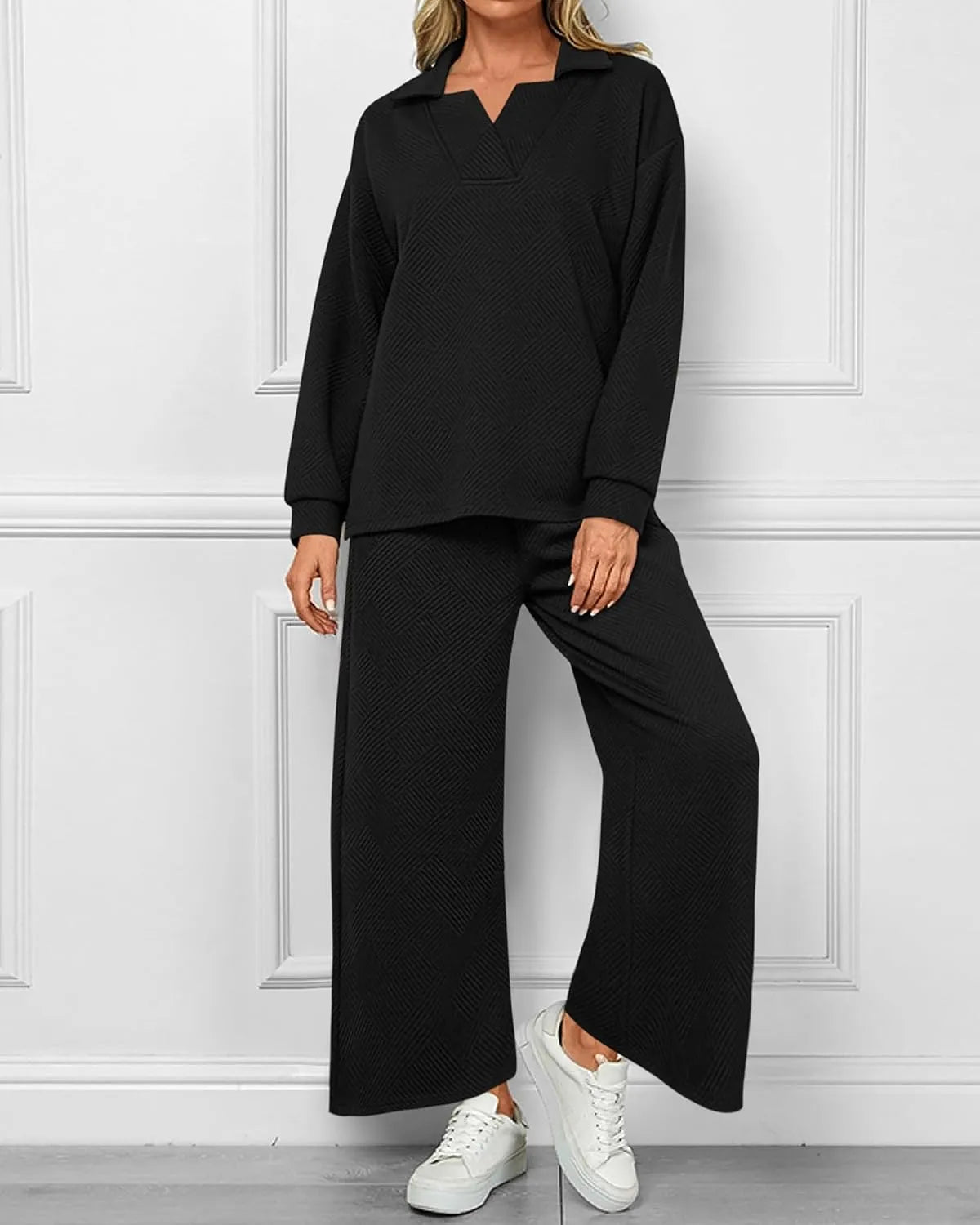 Person wearing a black outfit with wide-leg pants against a white paneled wall.