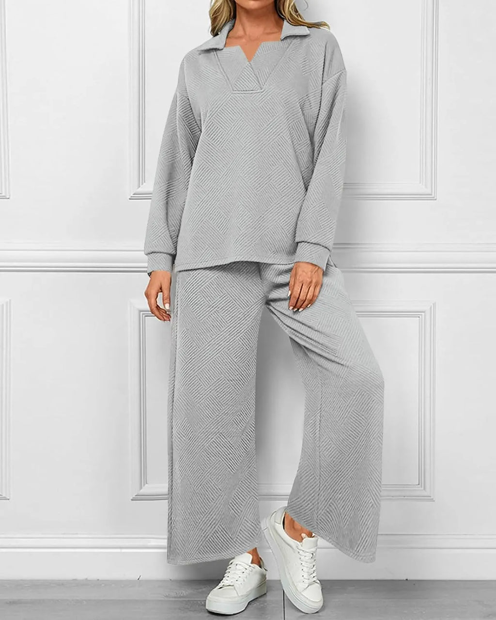 A woman models a light gray two-piece lounge set consisting of a long-sleeve collared top and matching wide-leg pants, paired with white sneakers, set against a white paneled wall.