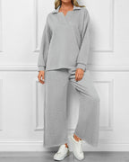 A woman models a light gray two-piece lounge set consisting of a long-sleeve collared top and matching wide-leg pants, paired with white sneakers, set against a white paneled wall.