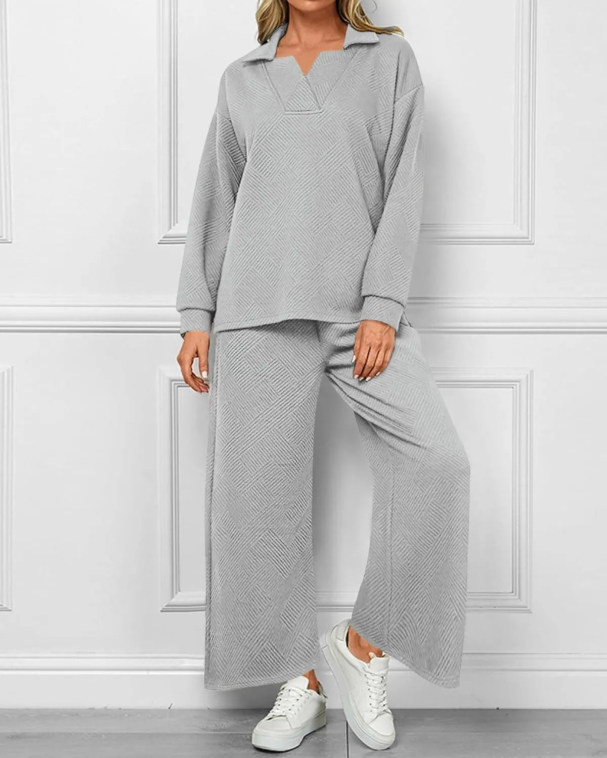 A woman models a light gray two-piece lounge set consisting of a long-sleeve collared top and matching wide-leg pants, paired with white sneakers, set against a white paneled wall.