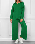 A woman modeling a matching green two-piece lounge set consisting of a long-sleeve collared top and wide-leg pants, paired with white sneakers, standing against a white paneled wall.